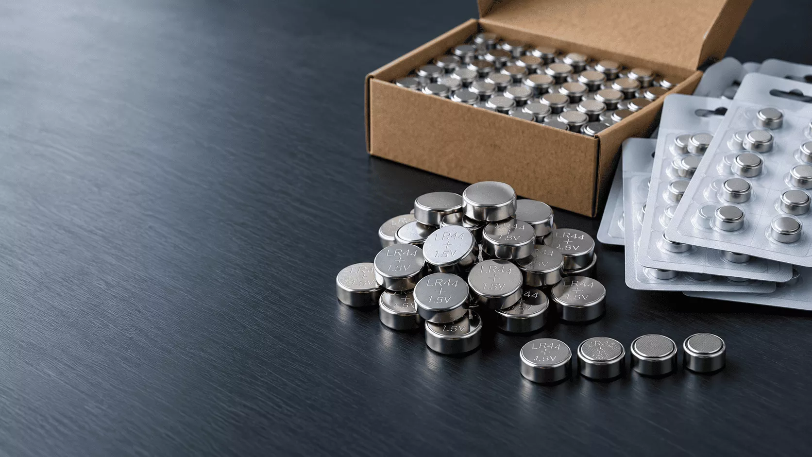 Pile of LR44 alkaline button cells beside a wholesale carton and blister packs on a dark slate surface
